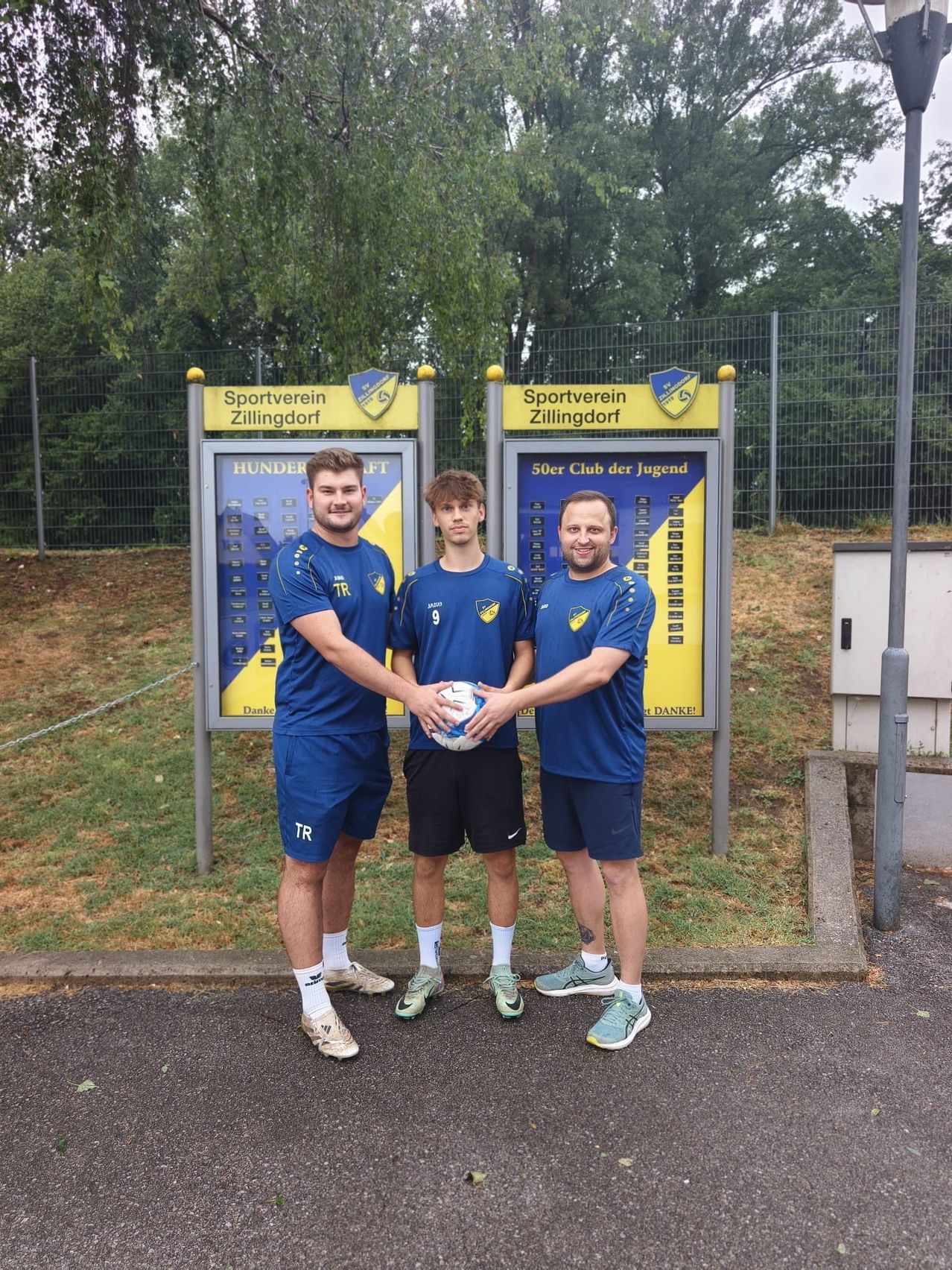 Drei Männer in blauen Uniformen stehen vor einem Schild des Sportvereins Zillingdorf. Der Mann in der Mitte hält einen Fußball.