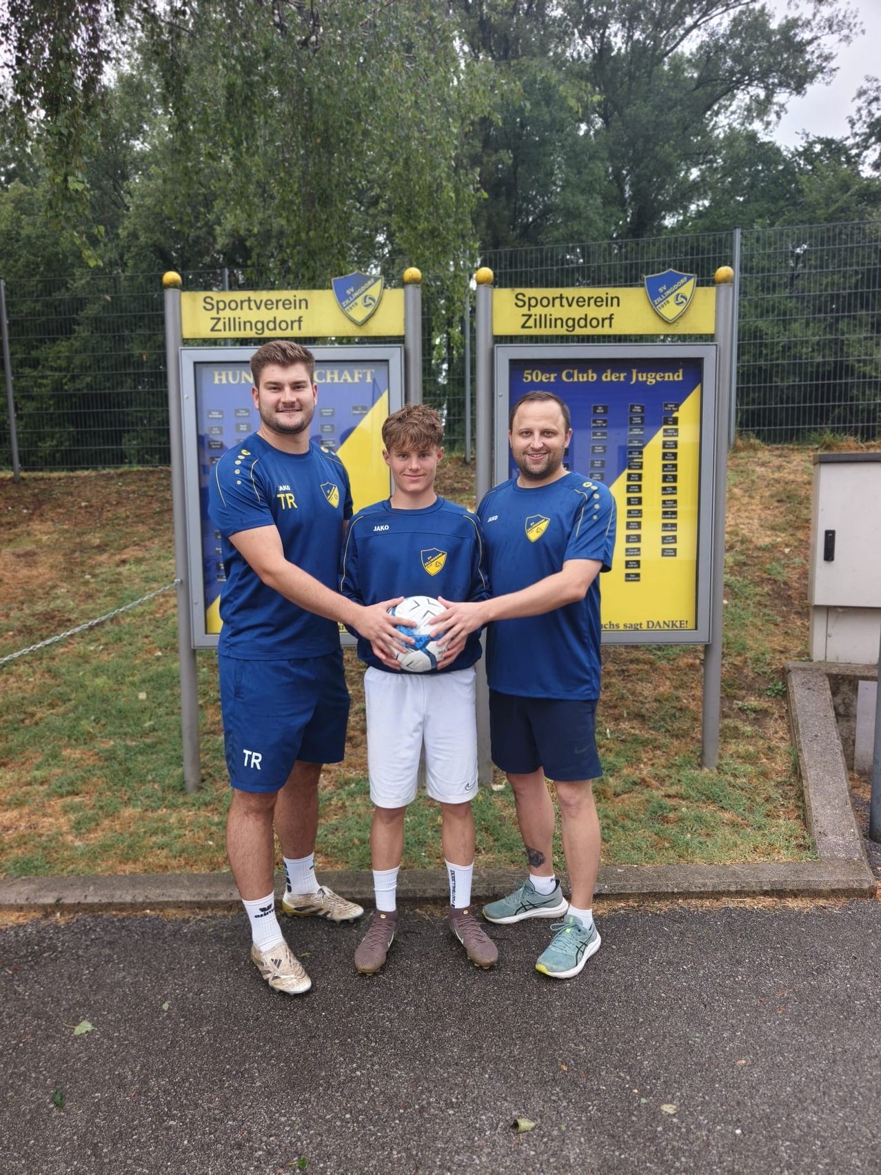 Drei Männer in blauen Trikots stehen vor einem Schild des Sportvereins Zillingdorf, einer hält einen Fußball.