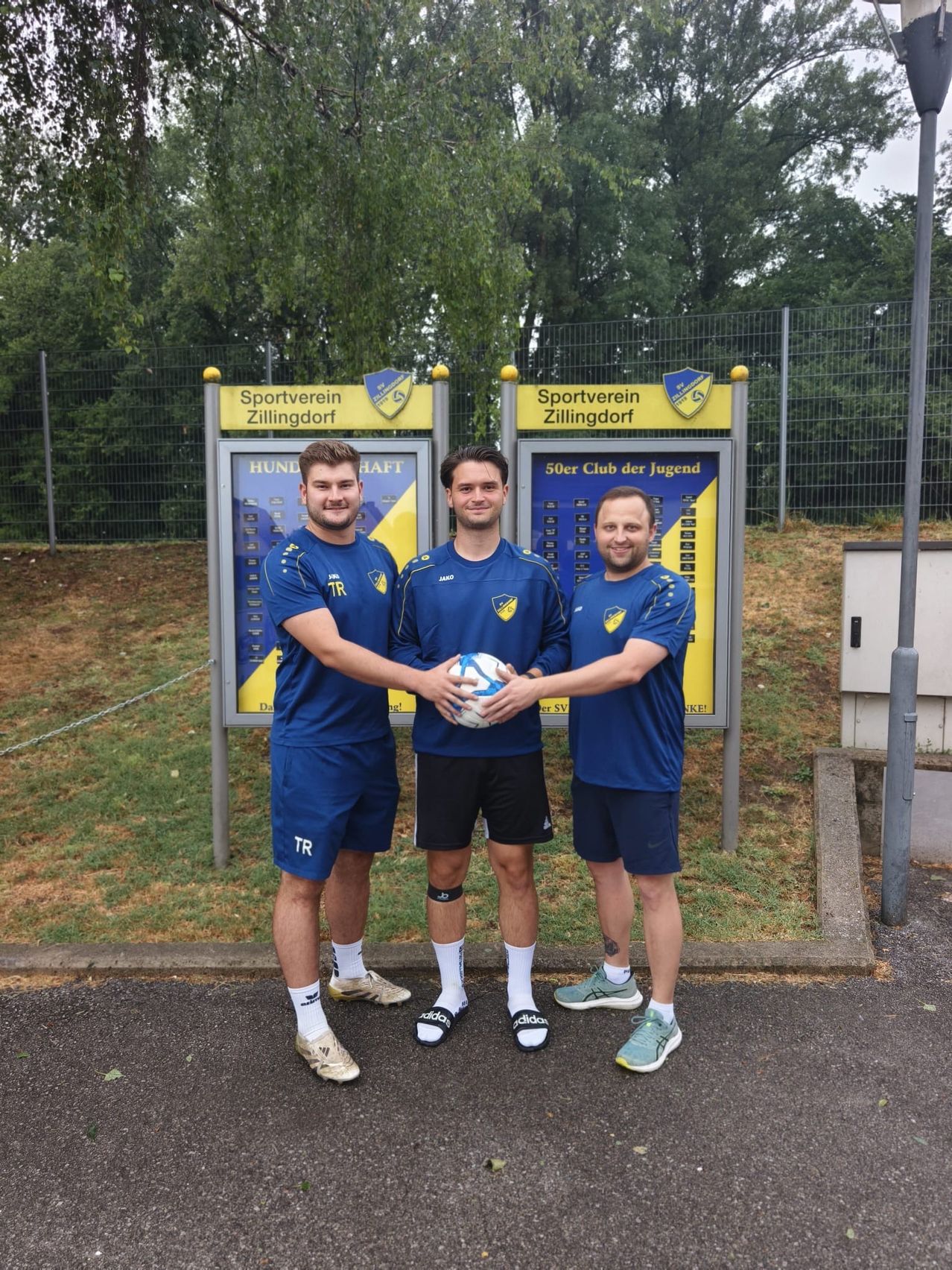 Drei Männer in blauen Uniformen stehen vor einem Schild des Sportvereins Zillingdorf und halten einen Fußball.