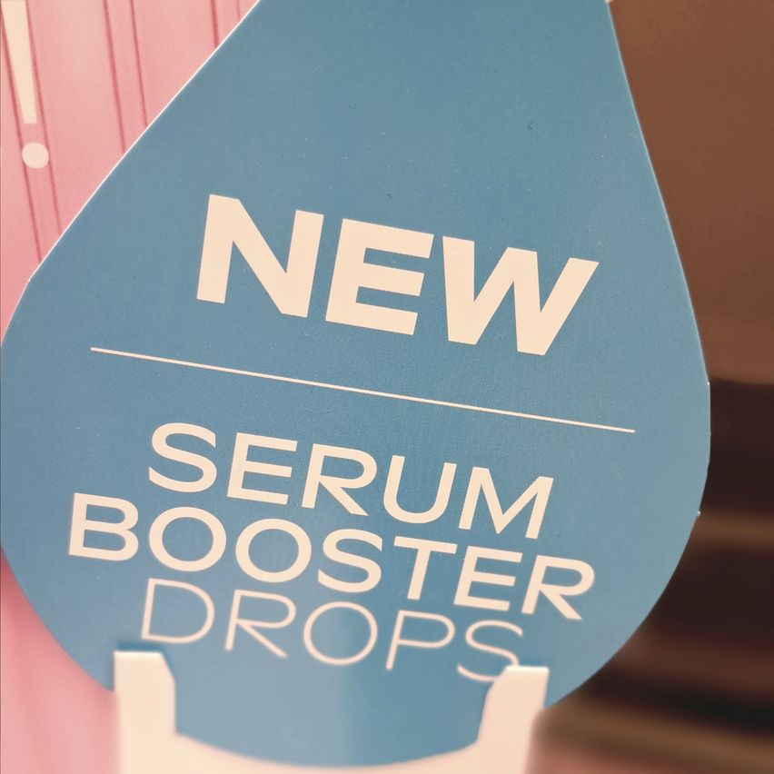 A blue sign in the shape of a droplet. It says 'NEW SERUM BOOSTER DROPS'. The background is pink.
