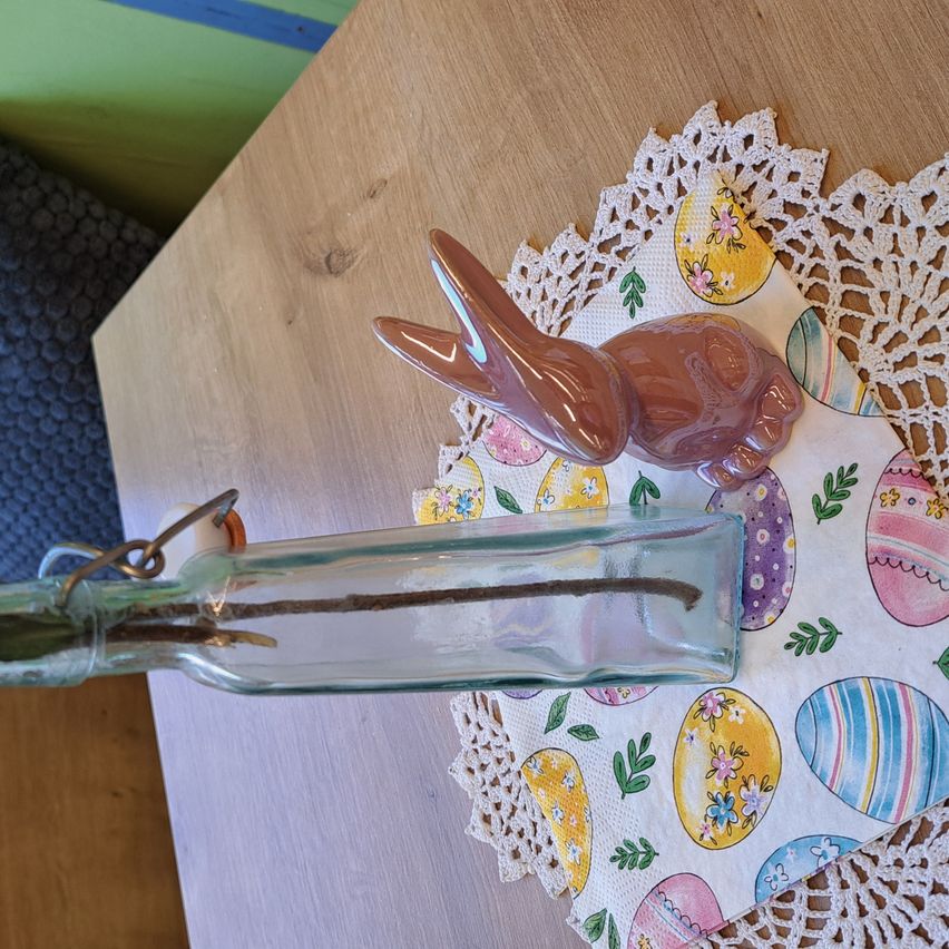 Eine durchsichtige Flasche mit einem Stock darin steht auf einem österlichen Doily. Ein brauner Keramikhase liegt auf der Flasche. Das Doily hat bunte Ostereier.