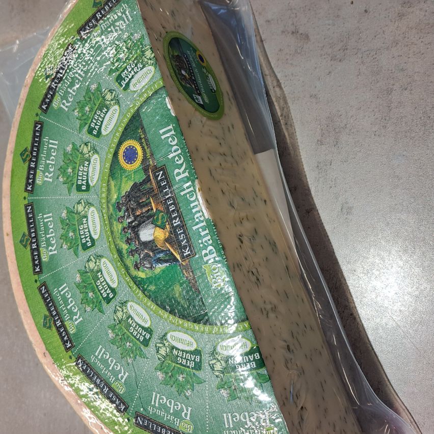 Ein Stück Käse, verpackt in Plastik mit einem grünen Etikett, das ein Design mit Menschen und einem Schloss zeigt. Das Etikett trägt 'BioRebel' und 'BioRebellen'.