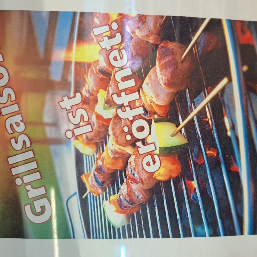 Ein Poster bewirbt Grillen und zeigt Spieße mit Fleisch und Gemüse auf einem Grill. Der Text lautet Grillspass ist ero�frei! in fetter Schrift. Der Hintergrund ist eine verschwommene Außenszene mit einem Zaun.