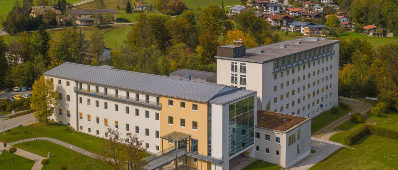 Luftaufnahme eines modernen Gebäudekomplexes mit Glasfassaden und einem grünen Dach, umgeben von Bäumen und einem grünen Feld im Hintergrund.