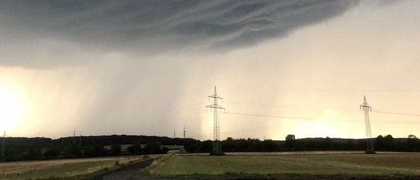 Ein bewölkter Himmel mit dicken Wolken über einer ländlichen Landschaft mit einem schmalen Weg und zwei hohen Stromleitungen.