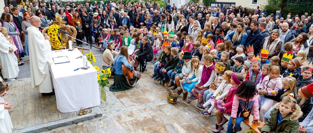 Eine große Menschenmenge, darunter Kinder mit Kronen, hat sich um eine zentrale Figur und einen Tisch mit einer Blumenarrangement versammelt. Die Veranstaltung scheint eine gemeinschaftliche Zusammenkunft oder Feier zu sein.