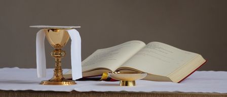 A gold chalice with a white cloth and an open book with a small bowl in front on a wooden table.