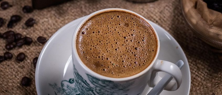 Eine Tasse Kaffee mit Löffel steht auf einer Untertasse. Im Hintergrund ist eine Schublade mit Kaffeebohnen und ein silberner Becher zu sehen.