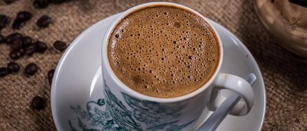 A cup of coffee sits on a saucer, surrounded by coffee beans. A metal container and wooden box filled with beans are in the background.