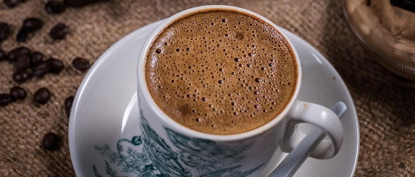 Eine Tasse Kaffee mit Schaum auf einer Untertasse mit einem Löffel. Ein Metallbecher mit Kaffeebohnen und eine Holzschublade sind im Hintergrund.