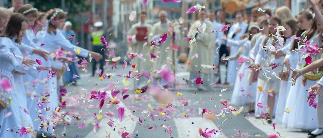 Eine Straßenszene mit Geistlichen, die gehen und Blütenblätter werfen. Die Straße ist mit bunten Blütenblättern bedeckt. Im Hintergrund sind Gebäude und eine Person mit Hut zu sehen.