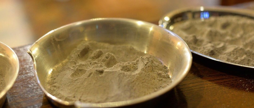 Ein metallischer Behälter mit feinem, hellbraunem Pulver steht auf einem Holztisch, wobei ein weiterer Behälter teilweise im Hintergrund zu sehen ist.