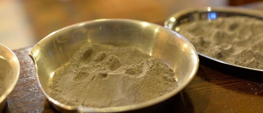 Ein metallischer Behälter mit feinem, hellbraunem Pulver steht auf einem Holztisch, wobei ein weiterer Behälter teilweise im Hintergrund zu sehen ist.