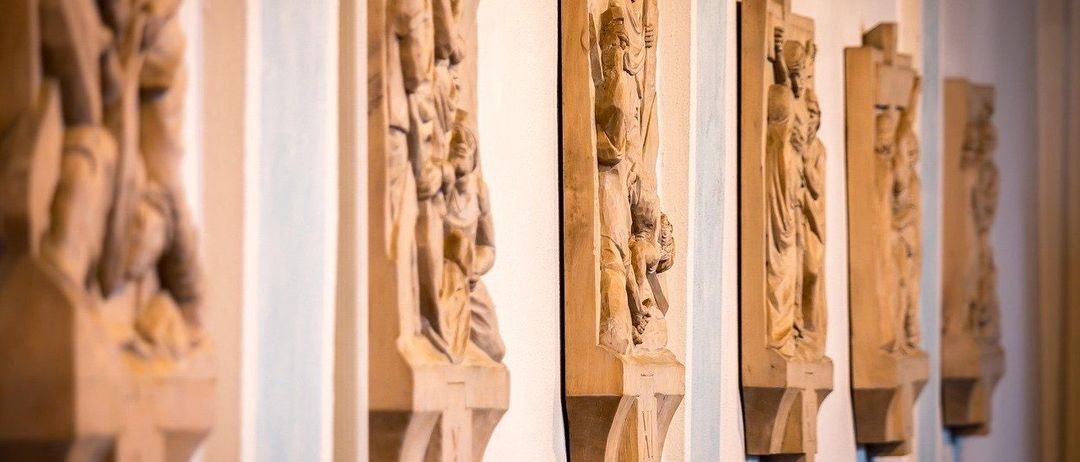 Eine Wand mit mehreren hölzernen Skulpturen, die biblische Szenen darstellen, jeweils in Hochrelief geschnitzt und an einer cremefarbenen Wand montiert.