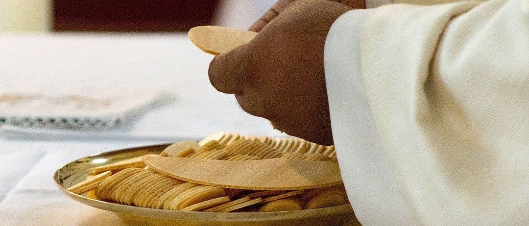 Eine Person in einem weißen Gewand legt einen Oblaten auf einen goldenen Teller, der mit weiteren Oblaten gefüllt ist, auf einer weißen Tischdecke.