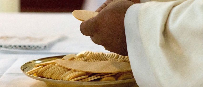 Eine Person in einem weißen Gewand legt einen Oblaten auf einen goldenen Teller, der mit weiteren Oblaten gefüllt ist, auf einer weißen Tischdecke.