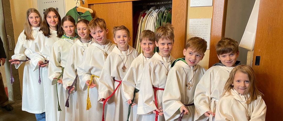 Eine Gruppe von Kindern in weißen Gewändern und roten Schärpen, die lächelnd für ein Foto in einer Kirche posieren.