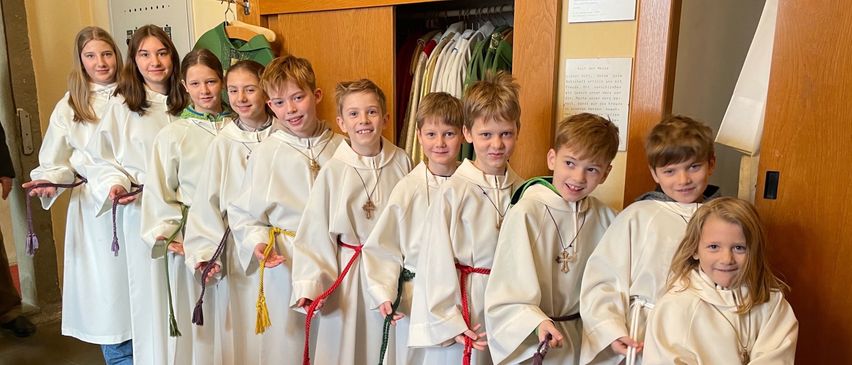 Eine Gruppe junger Kinder, alle in weißen Gewändern und roten Schärpen, posiert für ein Foto. Einige von ihnen halten bunte Bänder.