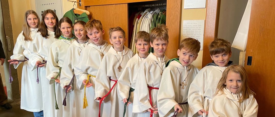 Eine Gruppe von Kindern in weißen Gewändern, wahrscheinlich Altardiener, steht in einem Raum zusammen. Der Raum ist mit einem hölzernen Schrank, weißen Wänden und einem roten Teppich ausgestattet.