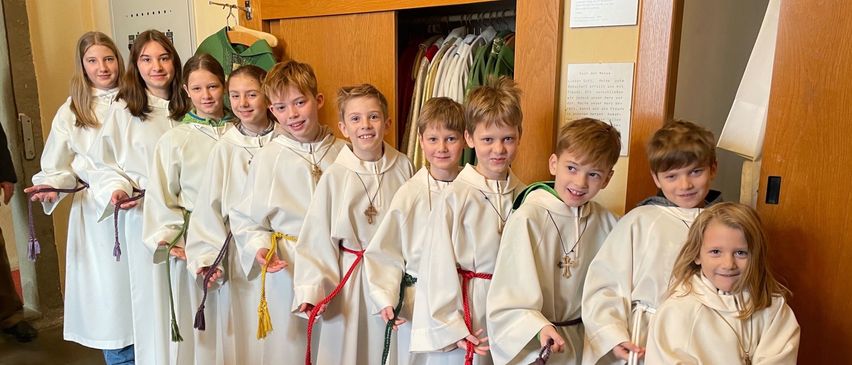 Eine Gruppe von Kindern in weißen Gewändern, wahrscheinlich Altardiener, steht in einem Raum zusammen. Der Raum ist mit einem hölzernen Schrank, weißen Wänden und einem roten Teppich ausgestattet.