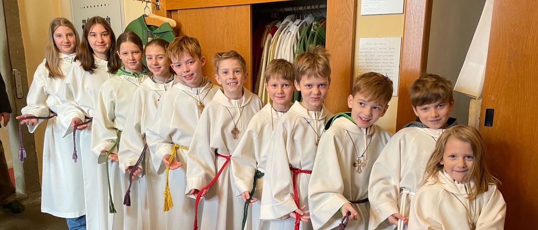 Eine Gruppe von Kindern in weißen Gewändern und roten Schärpen, die für ein Foto posieren. Sie halten ihre Schärpen und lächeln.