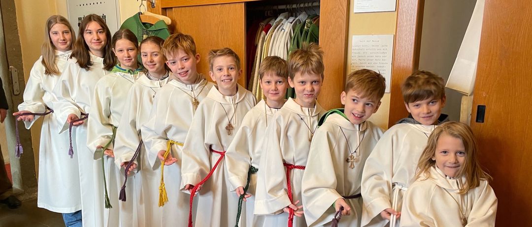 Eine Gruppe junger Kinder in weißen Gewändern mit roten Schärpen posiert für ein Foto in einem Raum.