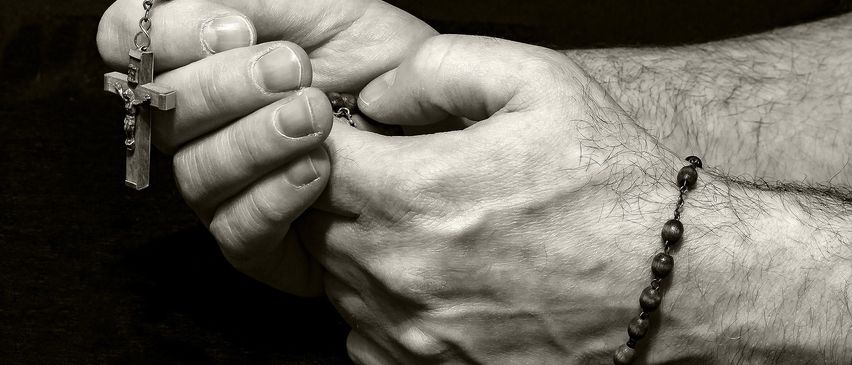 Nahaufnahme von zwei ineinander verschlungenen Händen mit einem Rosenkranz. Der Rosenkranz liegt über dem Arm der Person.