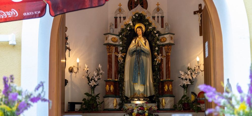 Eine Statue einer Frau mit einem Heiligenschein steht in der Mitte eines geschmückten Altars, umgeben von Blumen und Kerzen. Der Altar ist mit einem Kreuz und anderen religiösen Symbolen geschmückt.