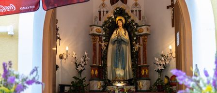 Eine Statue einer Frau mit einem Heiligenschein steht in der Mitte eines geschmückten Altars, umgeben von Blumen und Kerzen. Der Altar ist mit einem Kreuz und anderen religiösen Symbolen geschmückt.