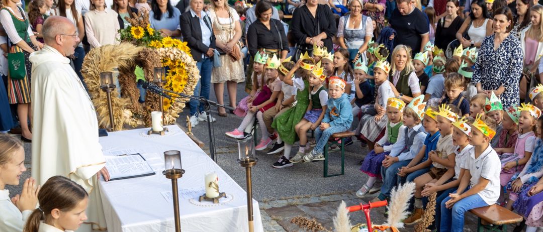 Eine große Menschenmenge, darunter Kinder mit Kronen, versammelt sich um einen Tisch mit einem Kranz und Kerzen, möglicherweise für eine religiöse Zeremonie.
