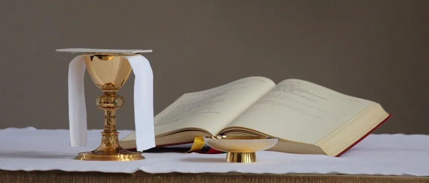 Ein goldener Kelch mit einem weißen Tuch und ein offenes Buch auf einem Tisch.