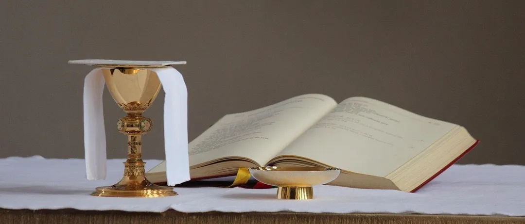 Ein goldener Kelch mit einem weißen Tuch und einem offenen Buch auf einem Tisch, möglicherweise für eine religiöse Zeremonie.