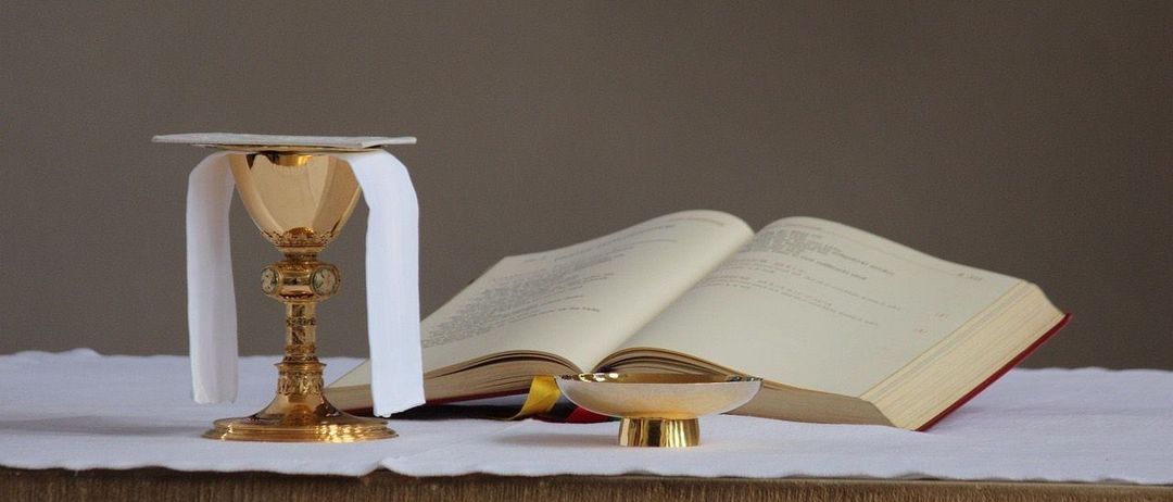 Ein goldener Kelch mit einem weißen Tuch und einem offenen Buch auf einem Tisch, möglicherweise für eine religiöse Zeremonie.