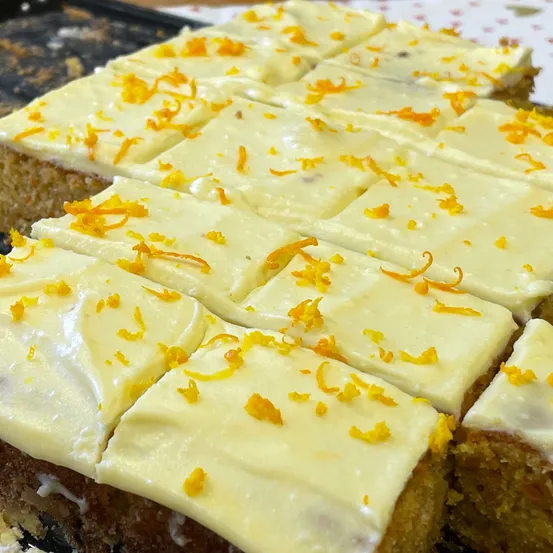 Ein Nahaufnahme eines geschnittenen Karottenkuchens mit weißer Glasur und Orangenschale. Jeder Kuchen ist großzügig mit Orangenschale belegt. Der Kuchen liegt auf einer schwarzen Oberfläche.