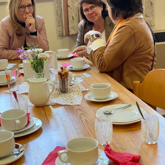 Drei Frauen sitzen um einen Tisch mit Tassen, Untertassen und einer Krug. Eine Frau hält eine Uhr und eine andere eine Karte. Ein Blumenvase mit Blumen steht auf dem Tisch.