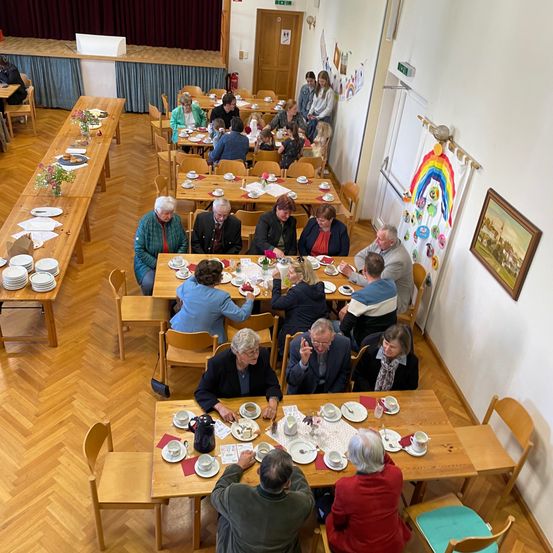 Eine Luftaufnahme eines Speisesaals mit vielen Menschen an Tischen. Es gibt Teller, Tassen und Blumen auf den Tischen. Ein Gemälde ist an der Wand.