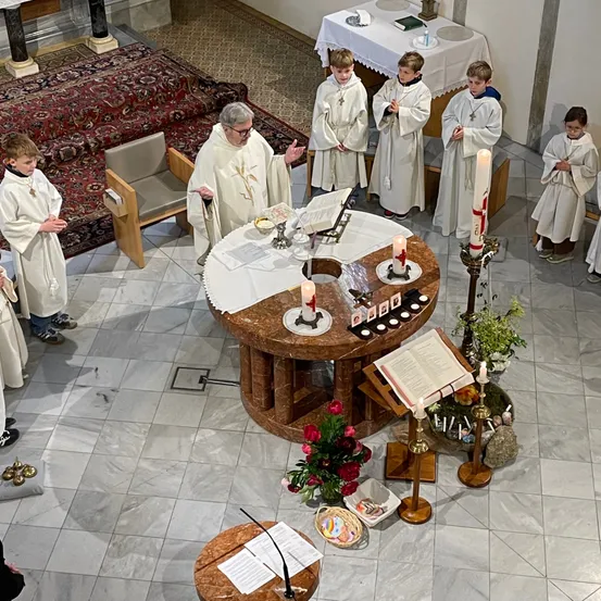 Ein Priester steht an einem Altar mit Kerzen. Junge Jungen in weißen Gewändern stehen um ihn herum. Der Altar hat Blumen, Kerzen und ein Buch. Im Hintergrund befinden sich ein Teppich, Stühle und ein Tisch.