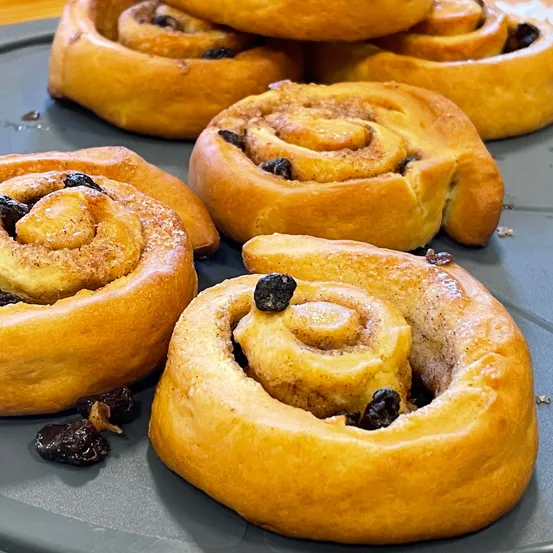 Mehrere goldbraune Zimtschnecken mit Rosinen sind auf einer grauen Oberfläche zu sehen. Jede Rolle hat ein Spiralmuster und einen Hauch von Glasur auf der Oberseite.