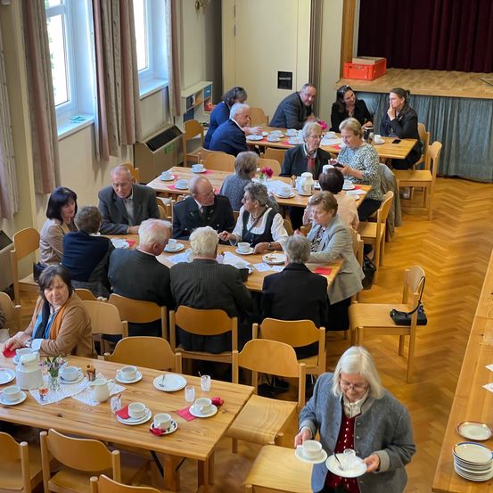 Mehrere Erwachsene versammeln sich in einem Esszimmer mit Holztischen und Stühlen. Sie essen mit Tassen und Untertassen. Der Raum hat Fenster mit Vorhängen, einen Holzboden und eine Bühne mit einem roten Kasten obenauf.