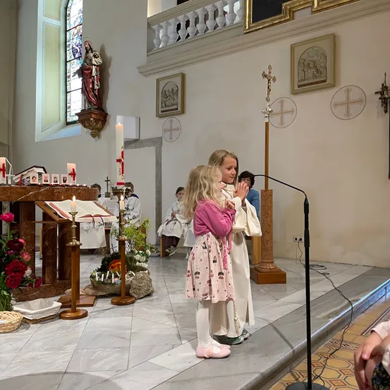 Zwei junge Mädchen stehen vor einem Mikrofon, möglicherweise in einer Kirche. Eine Frau hält die Hände eines Mädchens. Ein Kerzenständer mit brennenden Kerzen steht links. Eine Statue ist an der Wand über dem Kerzenständer angebracht.