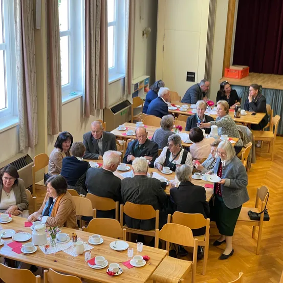 Eine Gruppe von Menschen sitzt um Tische in einem Raum und führt Gespräche, während sie ihre Mahlzeiten genießen. Der Raum hat Holzböden, Vorhänge und eine Bühne mit einem roten Vorhang.