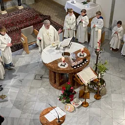 Ein Priester führt eine Zeremonie mit mehreren Kindern in weißen Gewändern durch. Der Altar ist mit Kerzen und Blumen geschmückt, und die Kinder beten.