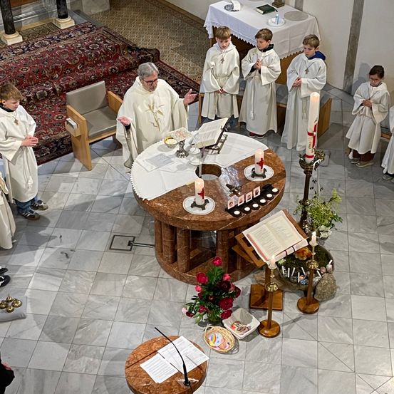 Ein Priester führt eine Zeremonie mit mehreren Kindern in weißen Gewändern durch. Der Altar ist mit Kerzen und Blumen geschmückt, und die Kinder beten.