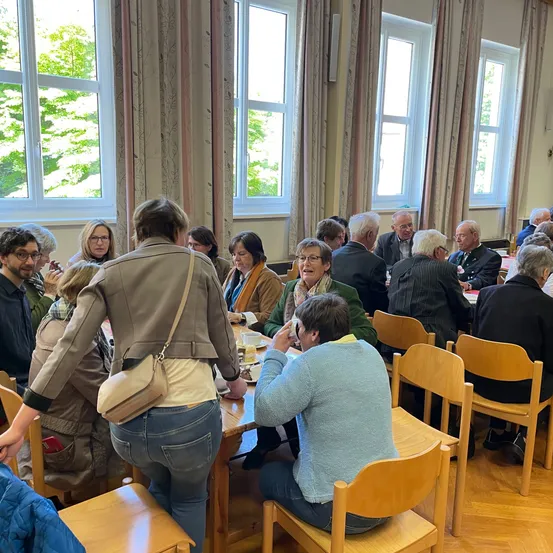 Eine Gruppe von Menschen sitzt um einen Tisch in einem Raum mit Holzböden, großen Fenstern und Vorhängen. Sie essen oder besprechen etwas.