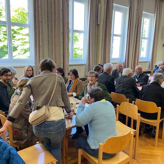 Eine Gruppe von Menschen sitzt um einen Tisch in einem Raum mit Holzböden, großen Fenstern und Vorhängen. Sie essen oder besprechen etwas.