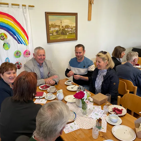 Eine Gruppe von Menschen sitzt um einen Tisch mit Tellern, Tassen und Blumen. Ein Mann und eine Frau lächeln und schauen in die Kamera. Hinter ihnen hängen ein Gemälde und ein Regenbogenbanner an der Wand.