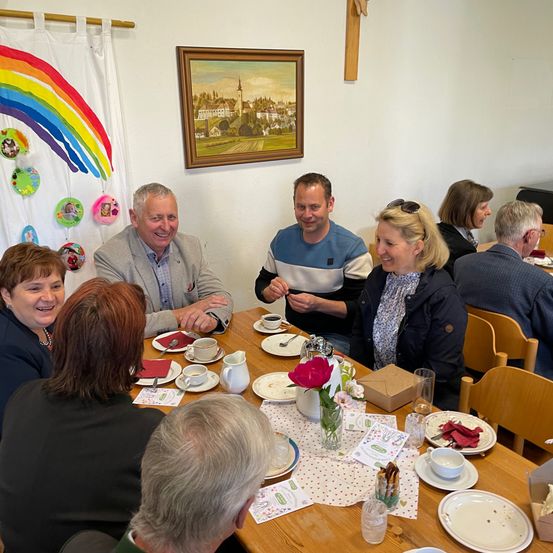 Eine Gruppe von Menschen sitzt um einen Tisch mit Tellern, Tassen und Blumen. Ein Mann und eine Frau lächeln und schauen in die Kamera. Hinter ihnen hängen ein Gemälde und ein Regenbogenbanner an der Wand.