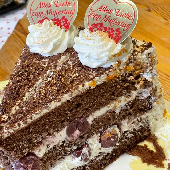 Ein Stück Schokoladenkuchen mit weißer Creme und Schokoladenraspeln, gekrönt von einem silbernen Herz mit der Aufschrift 'Alles Liebe zum Muttertag'. Er wird auf einem Teller auf einer Holzoberfläche serviert.