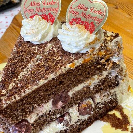 Ein Stück Schokoladenkuchen mit weißer Creme und Schokoladenraspeln, gekrönt von einem silbernen Herz mit der Aufschrift 'Alles Liebe zum Muttertag'. Er wird auf einem Teller auf einer Holzoberfläche serviert.