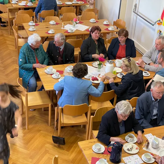 Eine Gruppe von Menschen sitzt um einen Tisch in einem Raum, wahrscheinlich einem Café. Sie unterhalten sich und scheinen ihre Mahlzeit zu genießen. Der Tisch ist mit Tassen, Untertassen und Tellern besetzt. Der Raum hat Holzböden und weiße Wände.